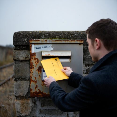 Ein Mann schiebt einen gelben Briefumschlag in einen verrosteten Briefkasten an einer maroden Bahnhofswand.