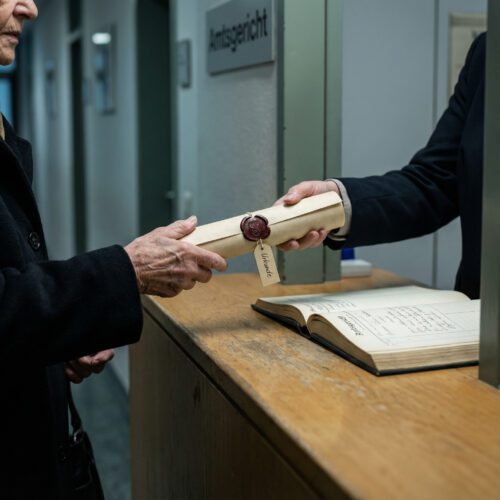 Ein Gerichtsbediensteter übergibt einer älteren Frau über einen schlichten Holztresen eine gerollte, gesiegelte Urkunde.