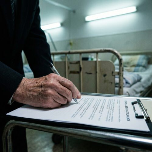 Nahaufnahme einer zittrigen Hand, die mühsam ein notarielles Dokument auf einem Klemmbrett im Krankenhaus unterzeichnet.