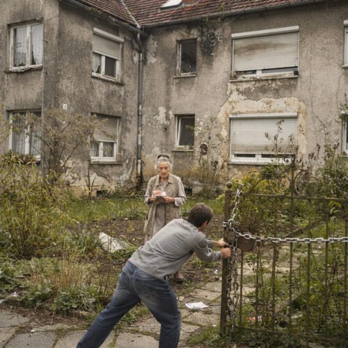 Hand einer Seniorin mit Geld vor feuchter Fassade, während ein Mann gegen das mit Ketten verschlossene Eisentor drückt.