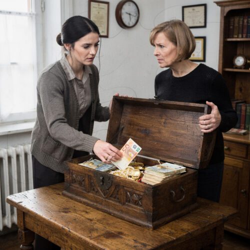 Eine Frau legt zögerlich einen dicken Geldstapel in eine antike Holztruhe, während eine zweite den schweren Deckel aufhält.