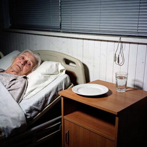 Eine ausgezehrte Seniorin blickt isoliert aus ihrem Pflegebett auf einen leeren Teller auf dem Nachttisch.
