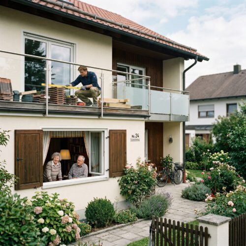 Ein Mann baut geschäftig das Obergeschoss um, während ein älteres Ehepaar hinter einem Fenster im Erdgeschoss steht.